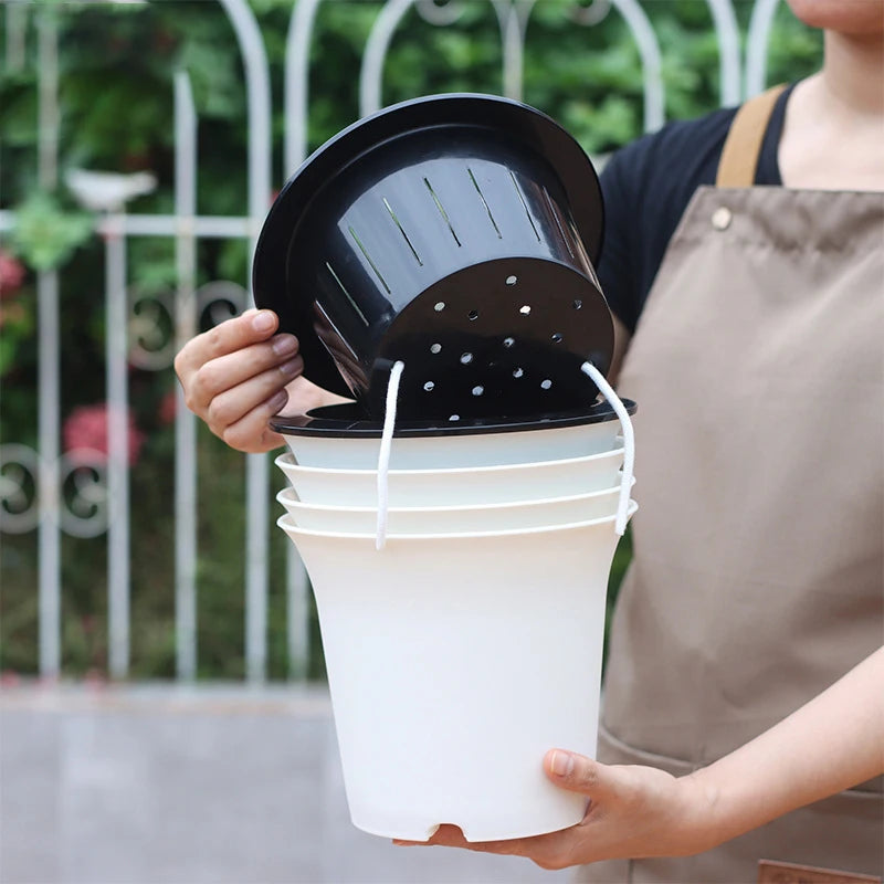 A person holds a white, self-watering flowerpot with a black inner basket featuring drainage holes and a cotton wick.