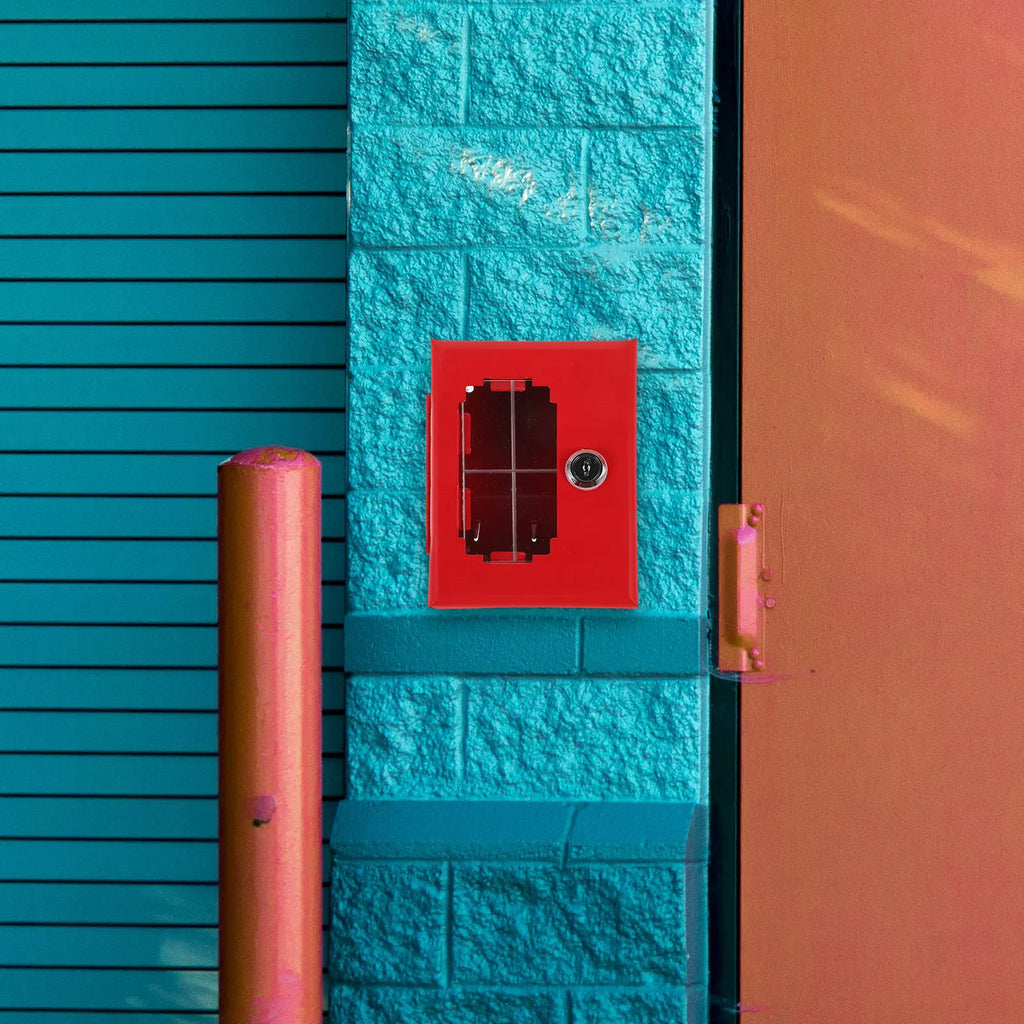 Red metal emergency key box with a lock, mounted on a textured teal brick wall next to a pink bollard and door.