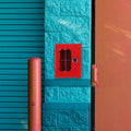 Red metal emergency key box with a lock, mounted on a textured teal brick wall next to a pink bollard and door.