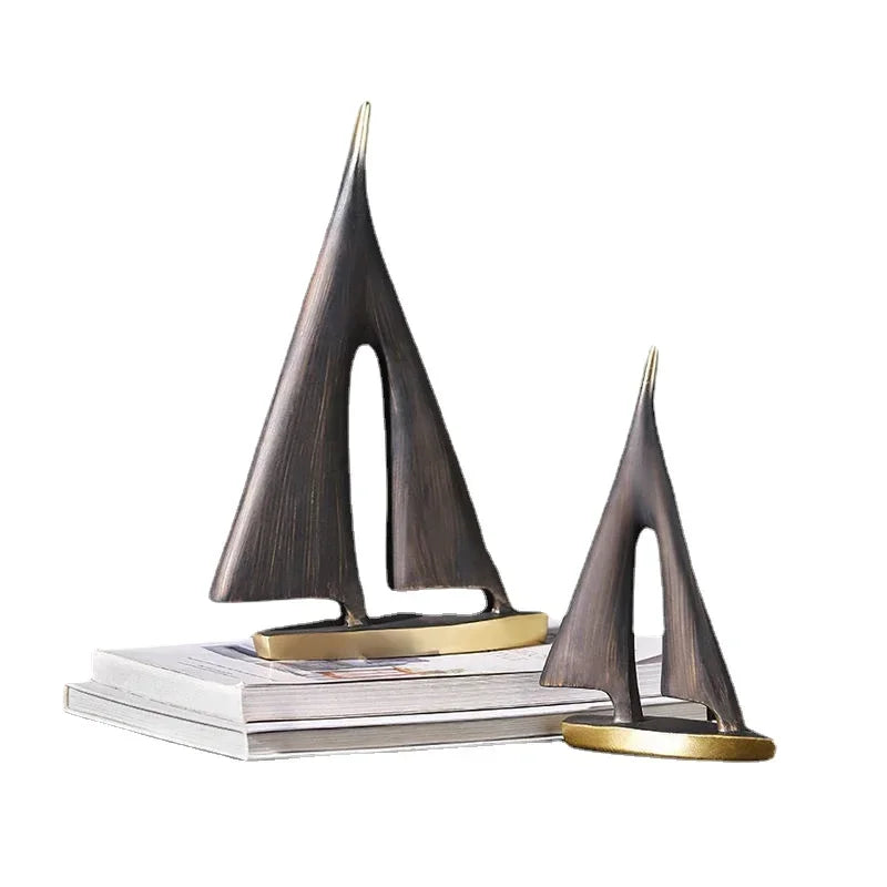 Two abstract, dark bronze-effect resin sailboat ornaments with gold bases displayed on a stack of books.