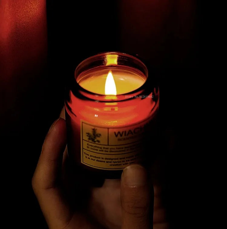 A hand holds a glowing scented candle in a glass jar, casting a warm, ambient light against a dark background.