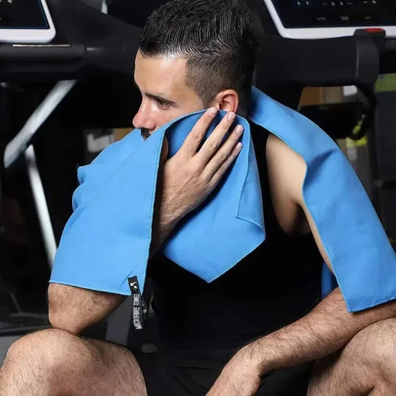 Man in a gym wiping his face with a bright blue microfibre towel featuring a black branded tab.