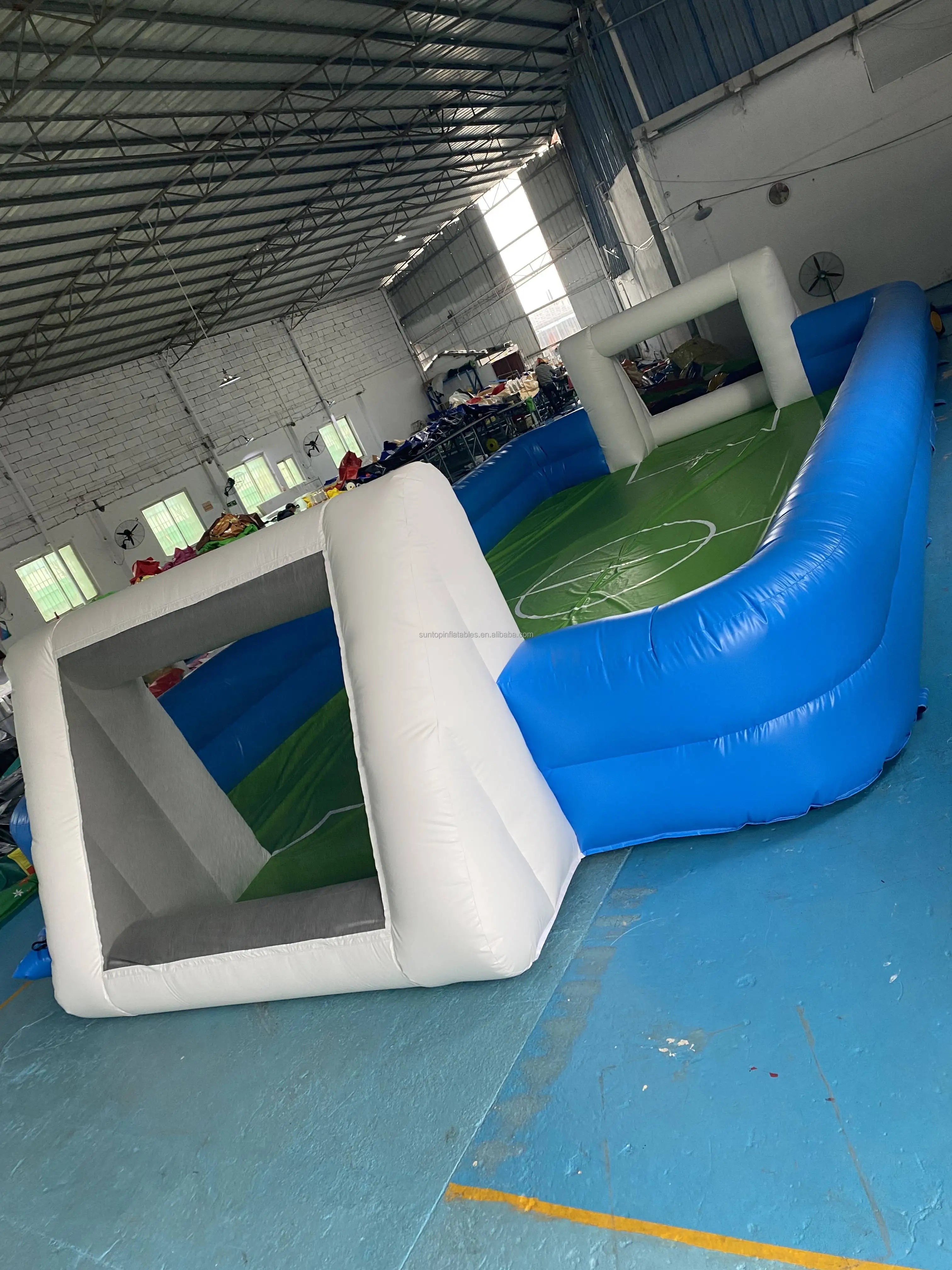 Blue and white inflatable football pitch with built-in goals and a green field marking, set up in a warehouse.