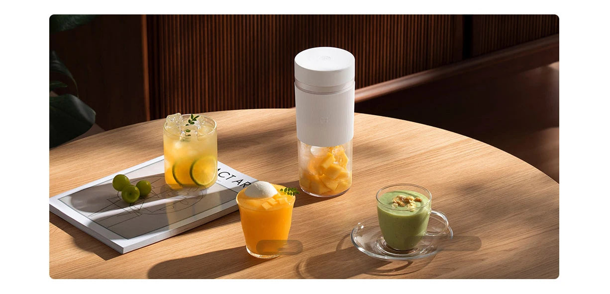A white portable blender on a wooden table, surrounded by three glasses containing colorful fruit juices and smoothies.