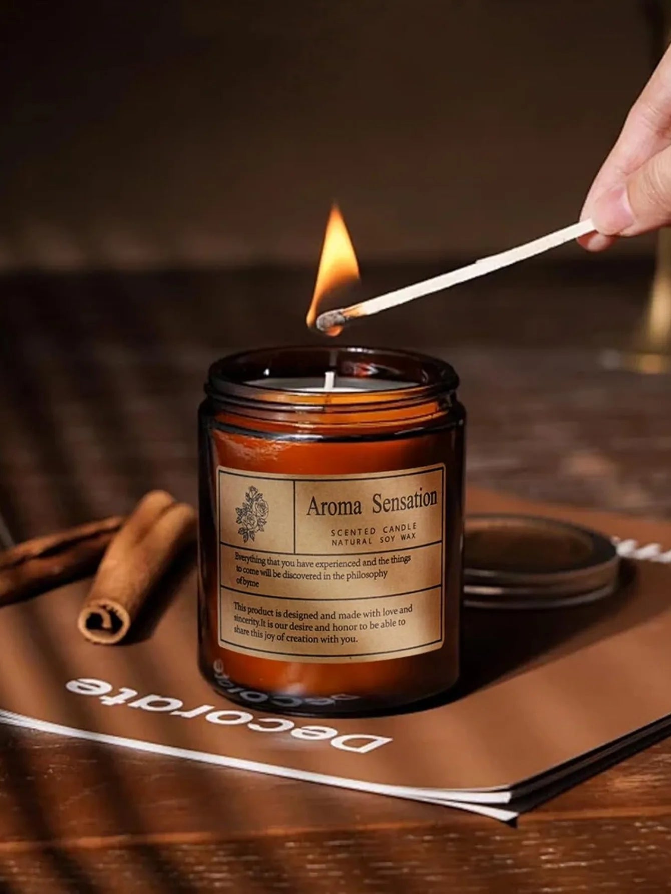 A hand lighting an amber-glass jar candle with a matchstick; natural soy wax candle on a rustic wooden table.