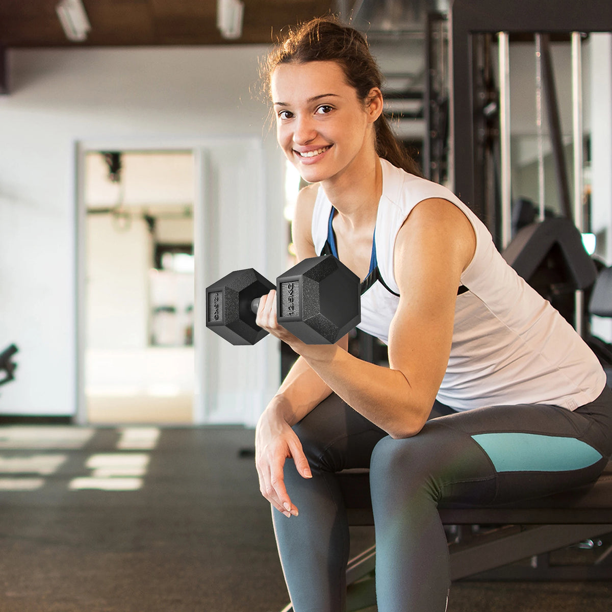Woman smiling while lifting a black 12.5kg rubber hex dumbbell in a gym.