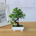 Artificial bonsai tree with green plastic leaves and a curved brown trunk in a small white rectangular pot.