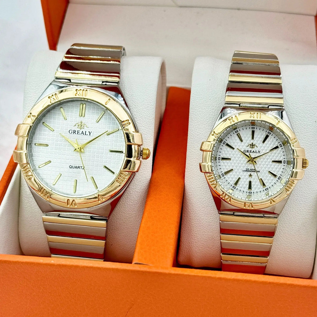 Two gold and silver quartz wristwatches with white textured dials and striped metal bands in an orange box.