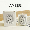 Amber scented candle in a glass votive with matching white gift box, featuring minimalist black oval typography.