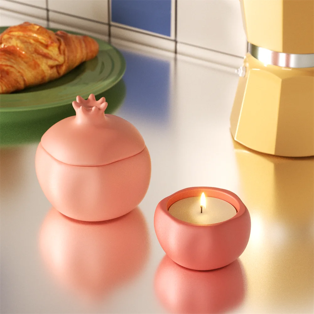 Two matte pink pomegranate candle vessels, one lidded jar and one lit tealight, on a metallic surface.