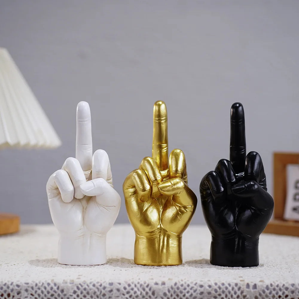 Three hand sculptures in white, gold, and black, each with a raised middle finger, standing on a textured surface.