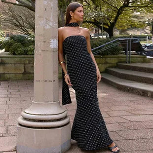 Black floor-length strapless column dress with white polka dots and a matching neck tie, worn outdoors.