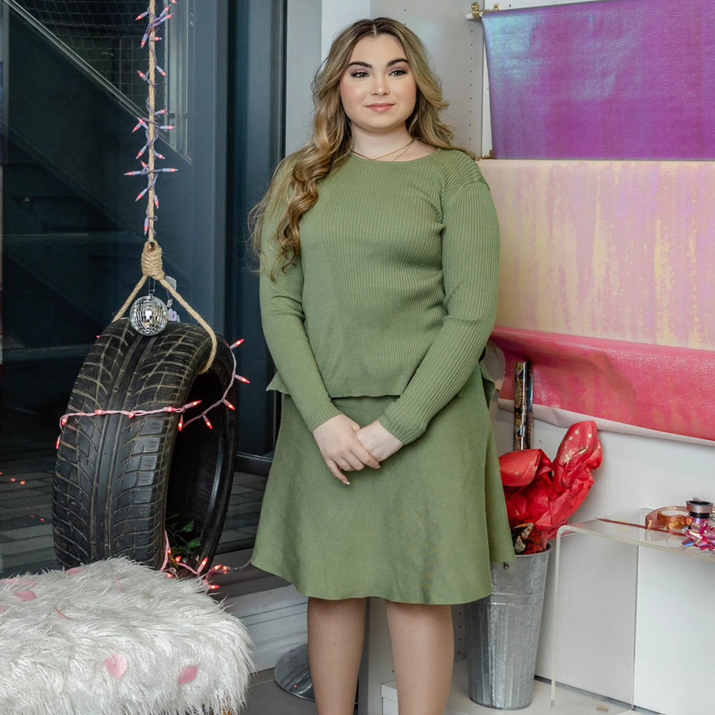 A woman wearing a sage green long-sleeved, ribbed-knit jumper paired with a matching flared midi skirt.