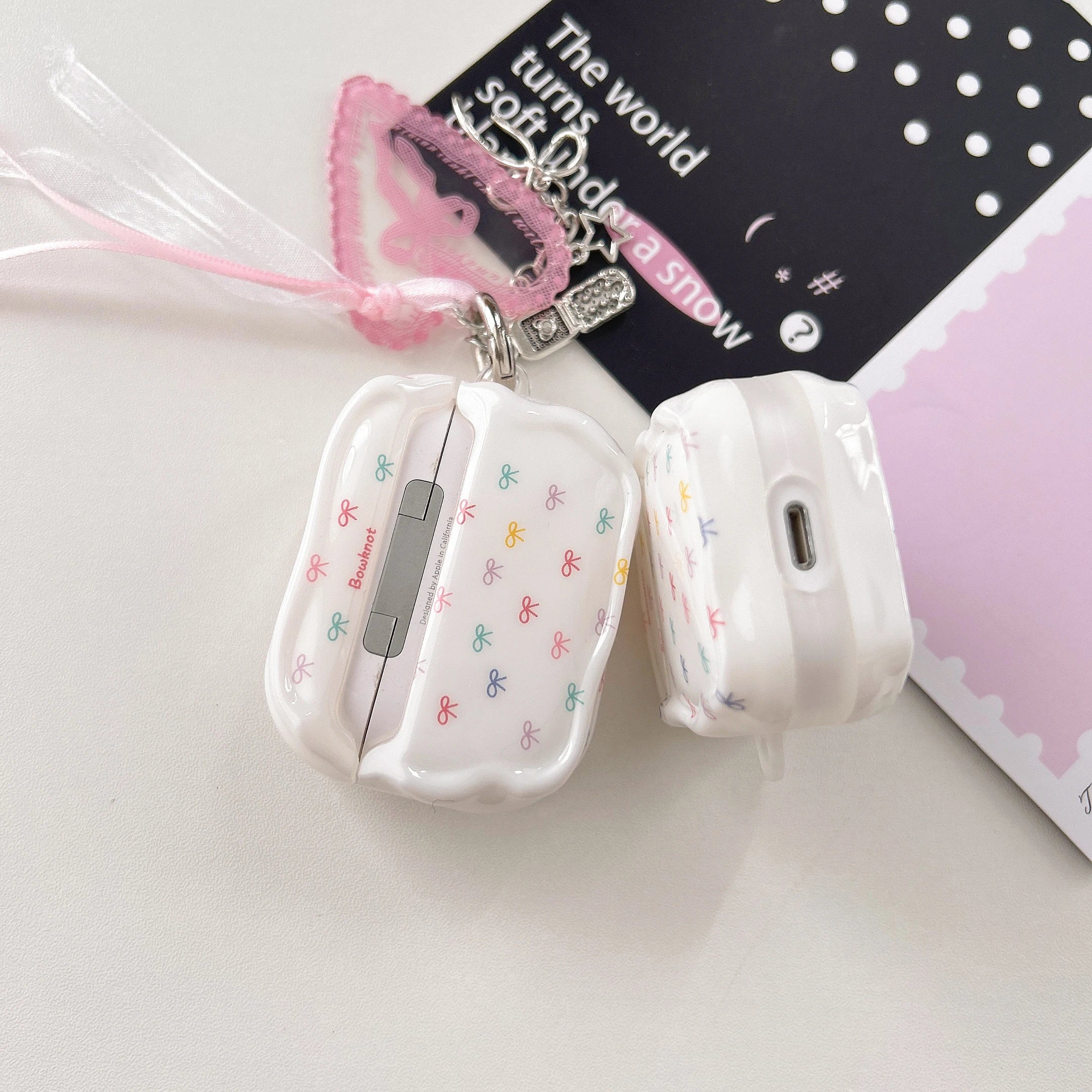 White glossy AirPods Pro case with colourful bow pattern and pink ribbon charm.