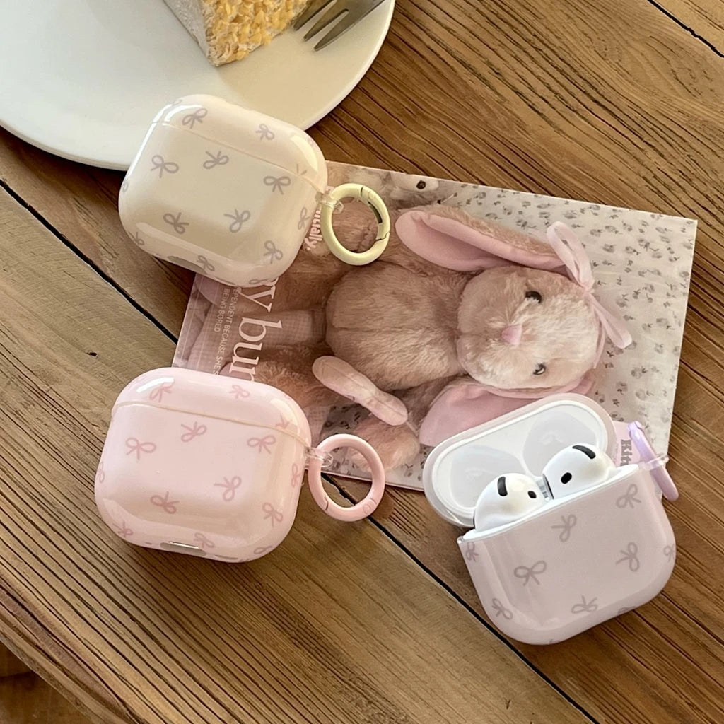 Cream and pink hard plastic AirPods cases with bow patterns and matching rings, compatible with Apple AirPods.