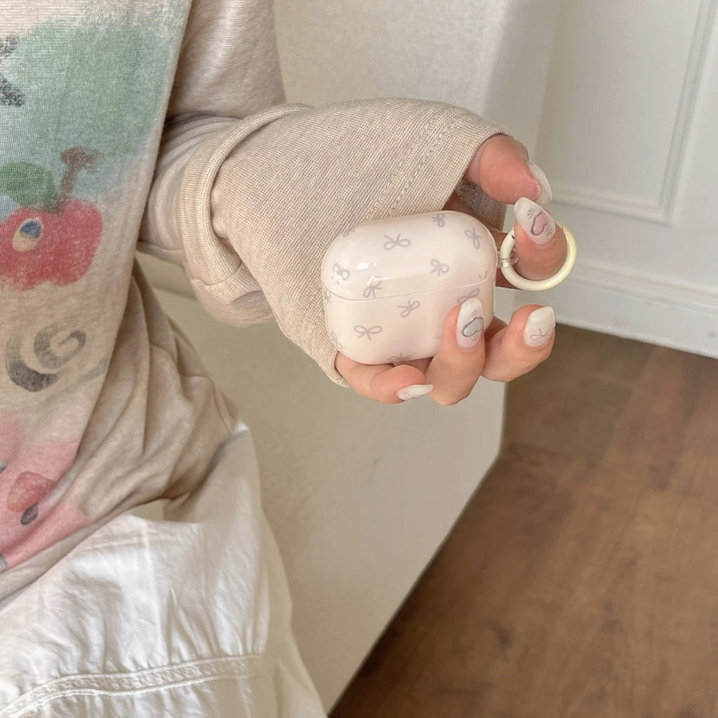 Pale pink AirPods Pro case with white bow print and matching ring, held in a hand.