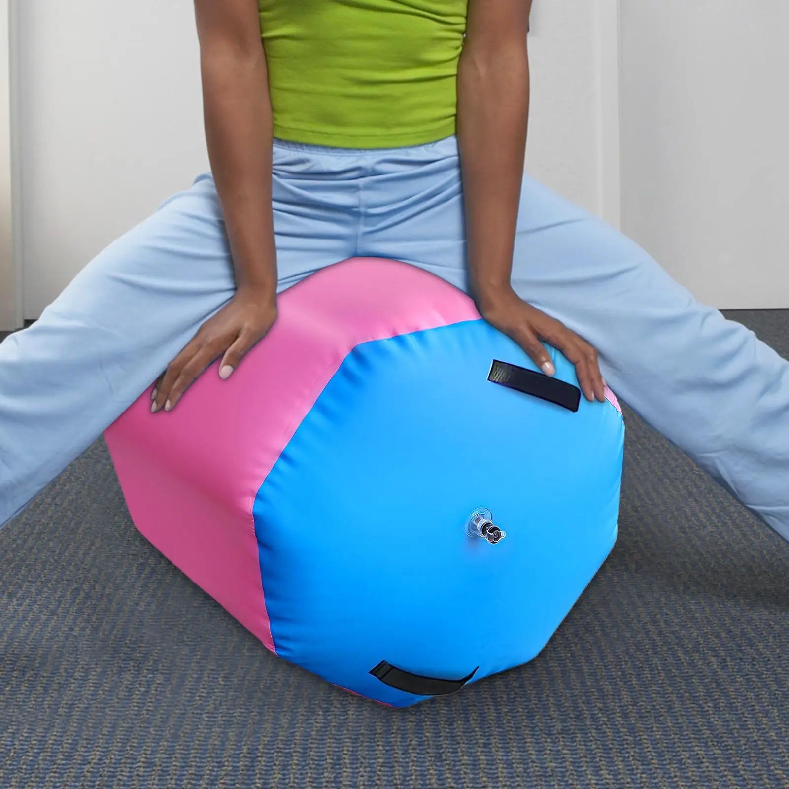 Person sitting on a pink and blue inflatable gymnastics air roller with black carry handles and an inflation valve.