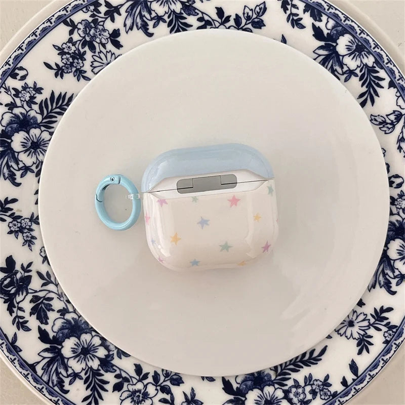 Glossy cream AirPods 3 case with pastel stars, a blue lid, and matching keyring, displayed on a patterned plate.