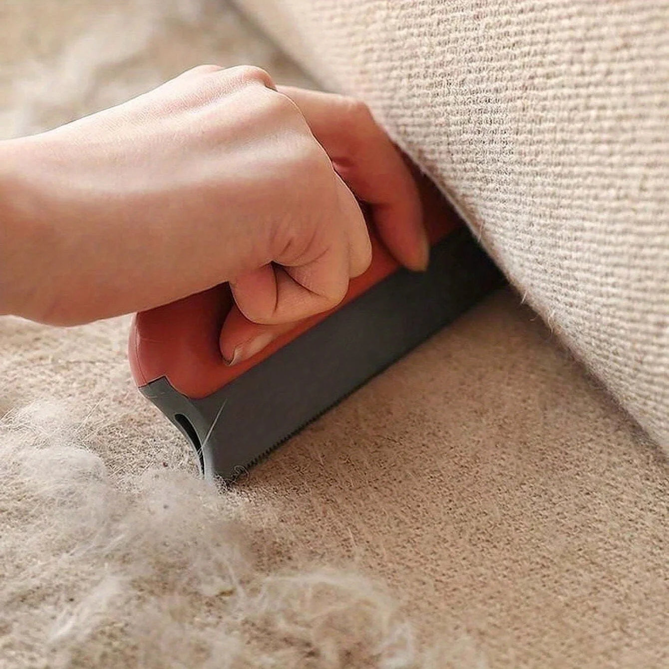 A hand using a red and grey handheld tool to scrape pet hair off a beige fabric sofa.