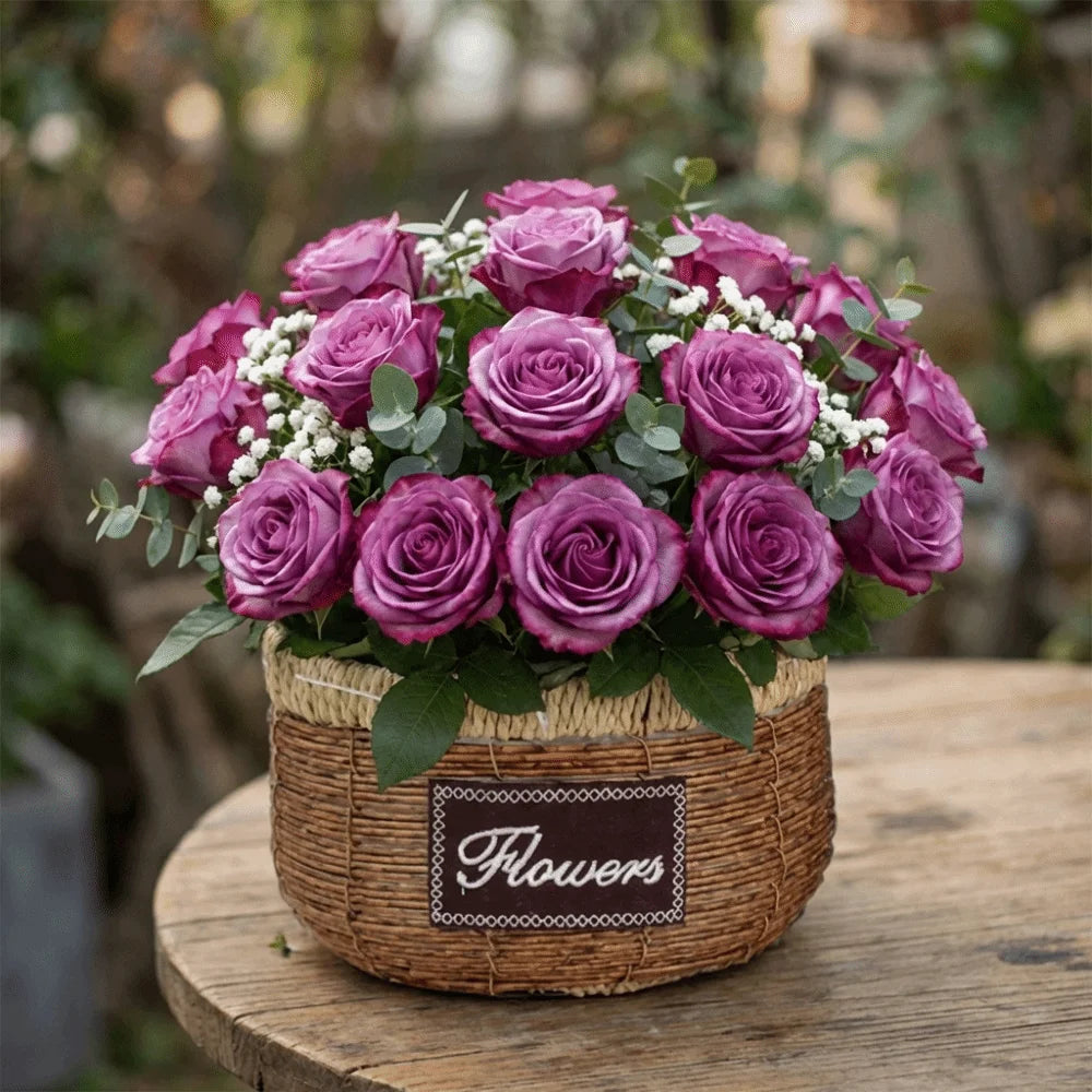 A rustic woven basket featuring a "Flowers" label, overflowing with purple roses and greenery on a wooden table.
