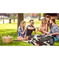 Cylindrical camouflage waterproof Bluetooth speaker sitting on the grass at a sunny park picnic.