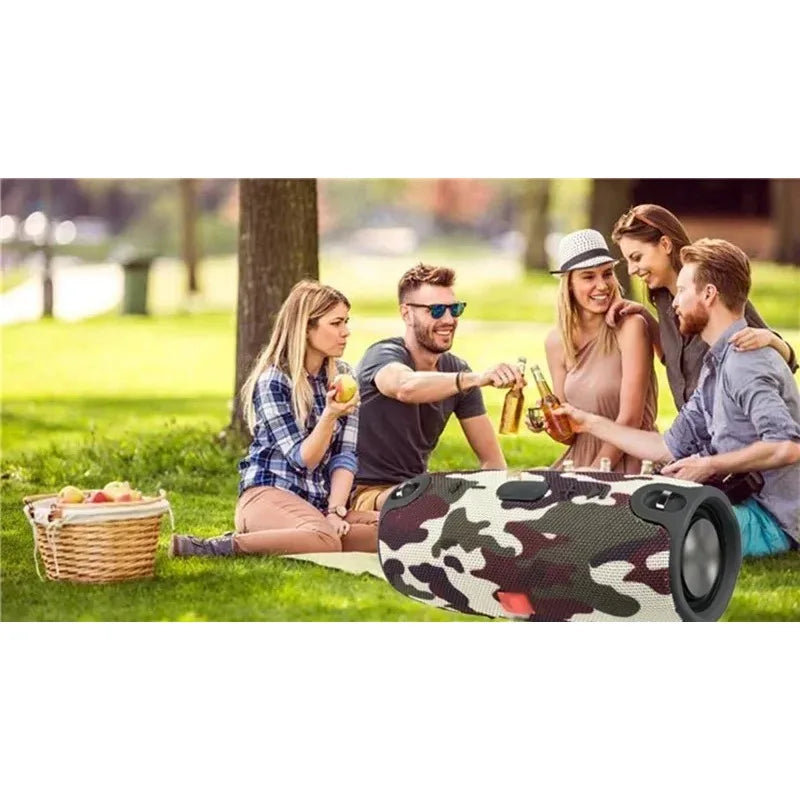 Cylindrical camouflage waterproof Bluetooth speaker sitting on the grass at a sunny park picnic.