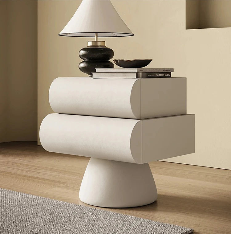 Cream modern bedside table with two rounded drawers on a pedestal base, styled with a lamp and books.