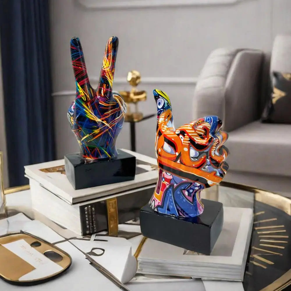 Two colourful, graffiti-style hand sculptures on black stands: one displaying a peace sign and one a thumbs-up gesture.