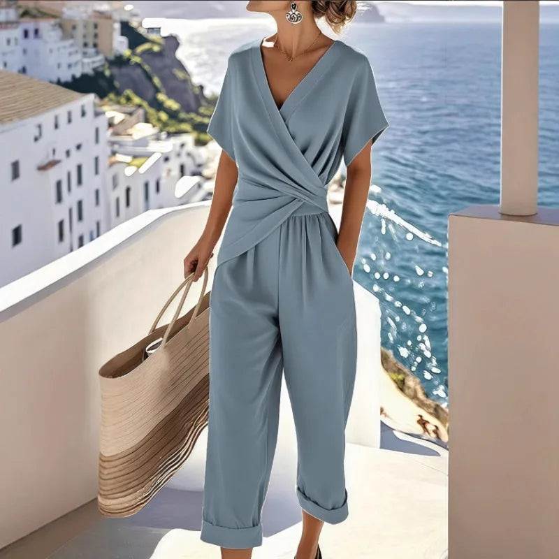 Dusty blue V-neck jumpsuit featuring short sleeves, a wrapped waist detail, and cuffed cropped trousers.