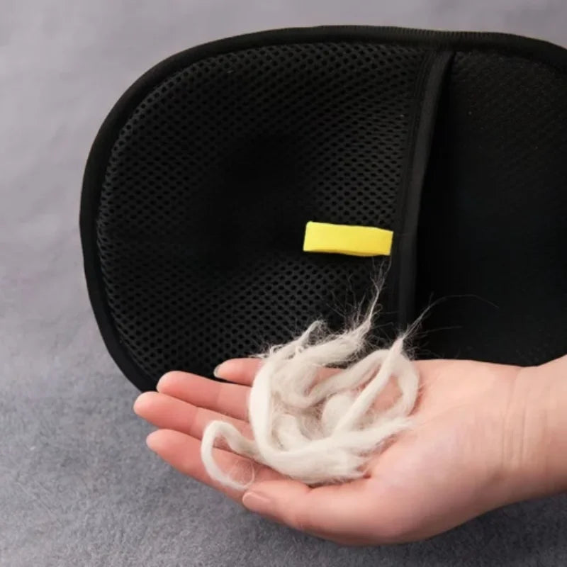 A hand holding a clump of white pet hair in front of a black, mesh grooming mitt with a yellow tab.