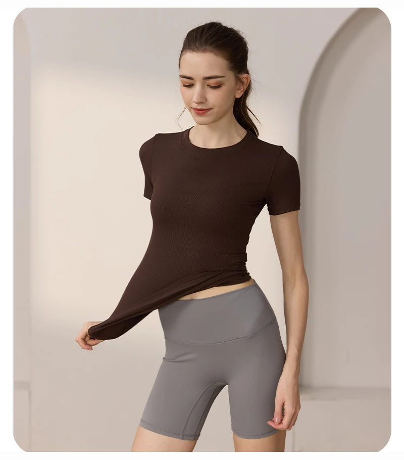 A woman wearing a dark brown, short-sleeved, ribbed crew-neck sports top with high-waisted grey athletic shorts.