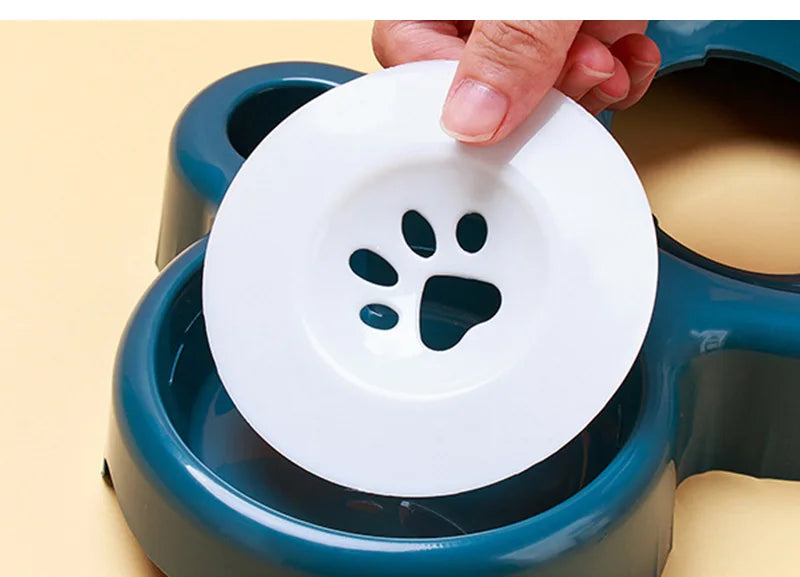 A hand placing a white, paw-print patterned floating disc into a dark teal, multi-section pet bowl.