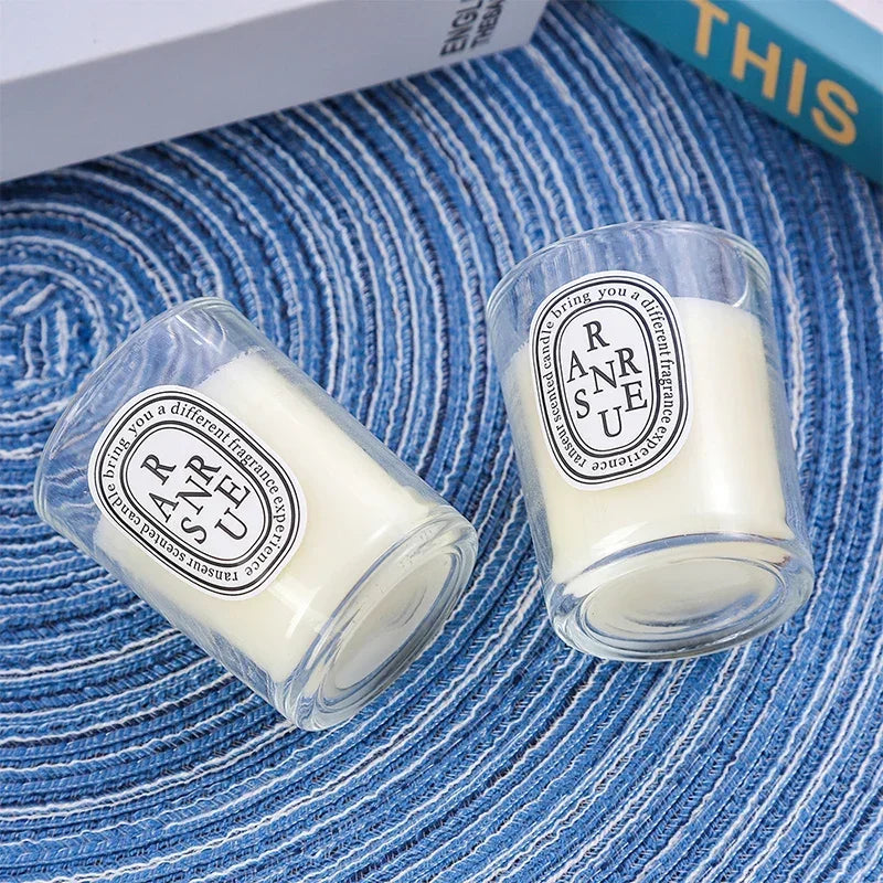Two small, clear glass votive candles with cream-coloured wax and minimalist oval labels on a blue textured mat.