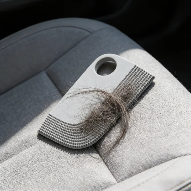 Grey silicone pet hair remover tool with a textured edge and finger hole, covered in collected hair on a car seat.