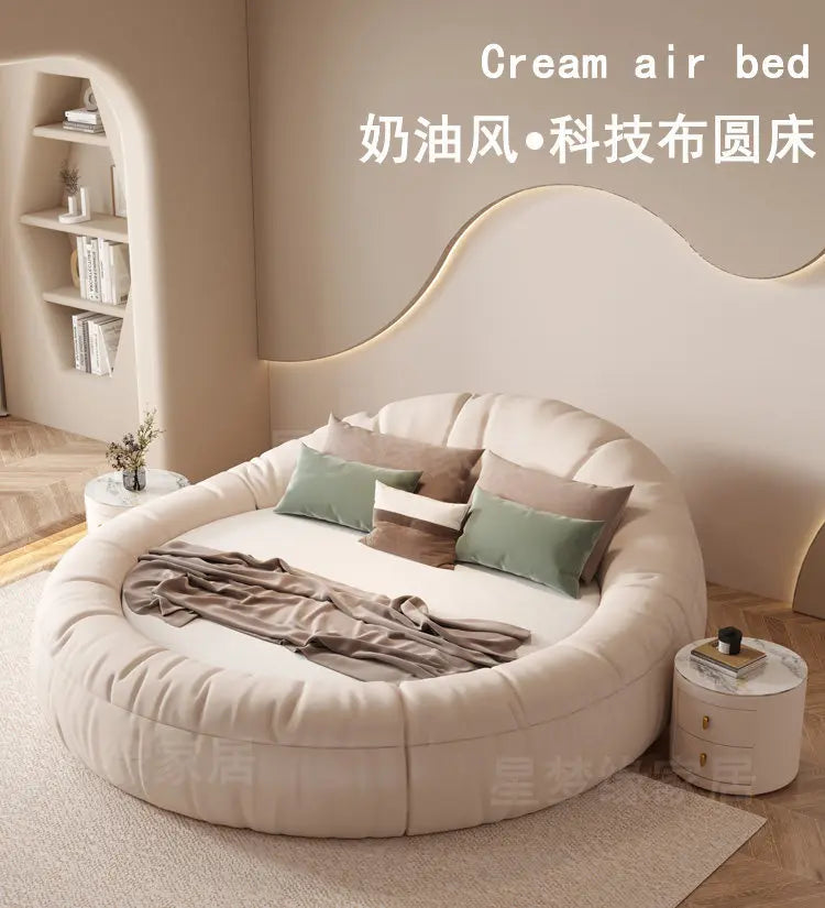 A modern, circular cream-coloured bed frame with a soft, padded finish, styled with cushions in a minimalist bedroom.