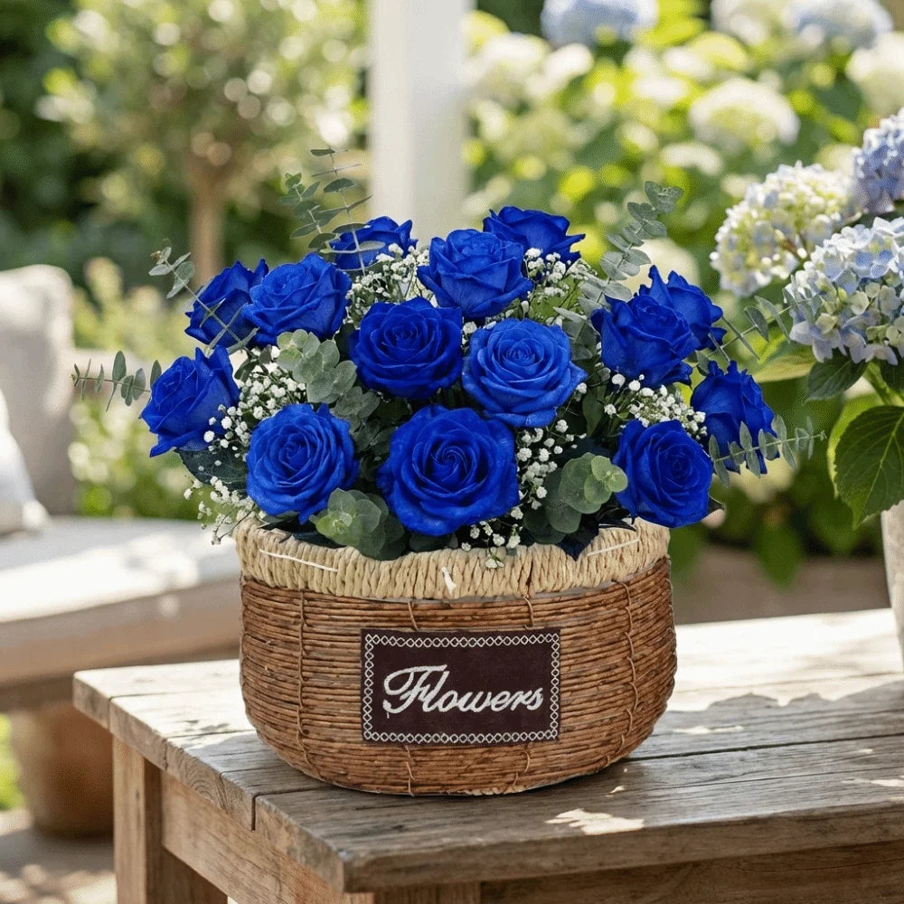 Vibrant blue roses with greenery arranged in a rustic, round woven basket featuring a "Flowers" label on a table.