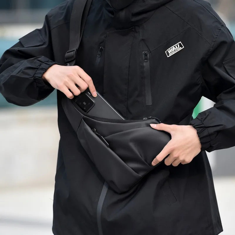 A sleek black, triangular anti-theft crossbody chest bag with zip pockets, worn over a black jacket.