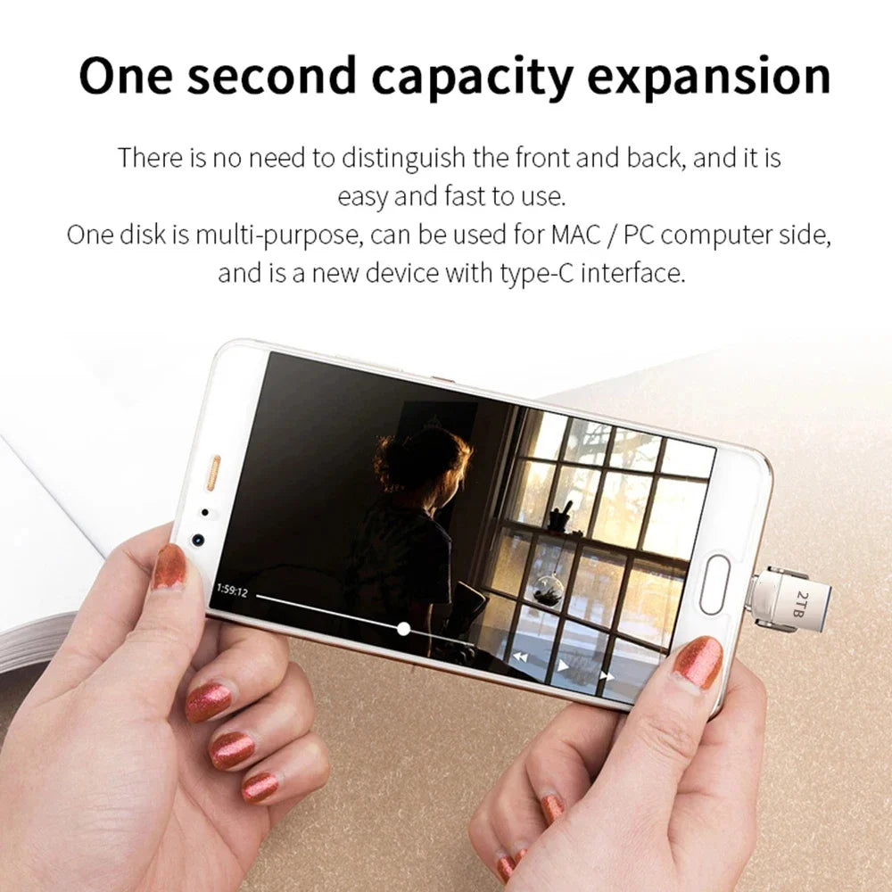 A person holds a smartphone with a compact, silver 2TB metal USB Type-C flash drive plugged into the charging port.