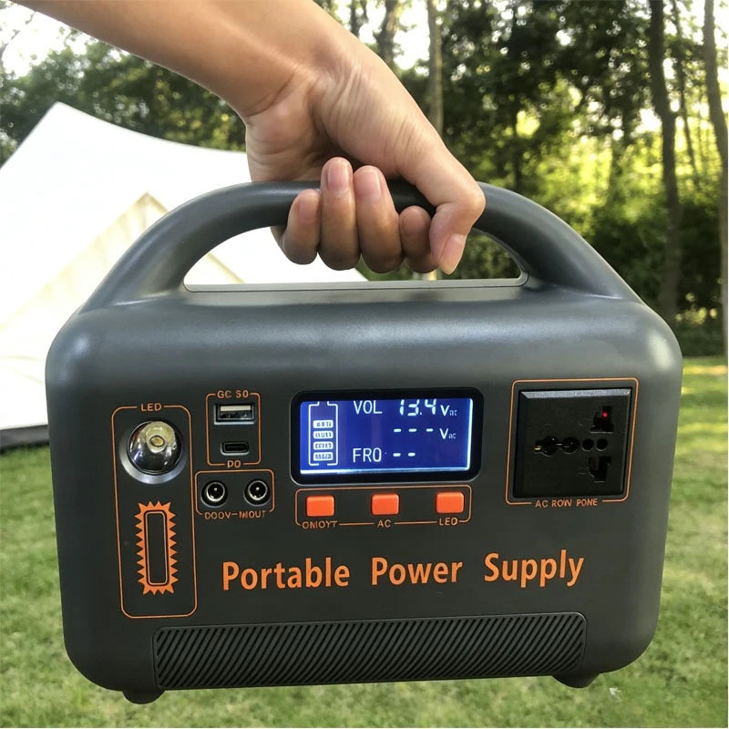 A hand holds a gray portable power supply with an LED screen, AC outlet, and USB ports in a campground setting.