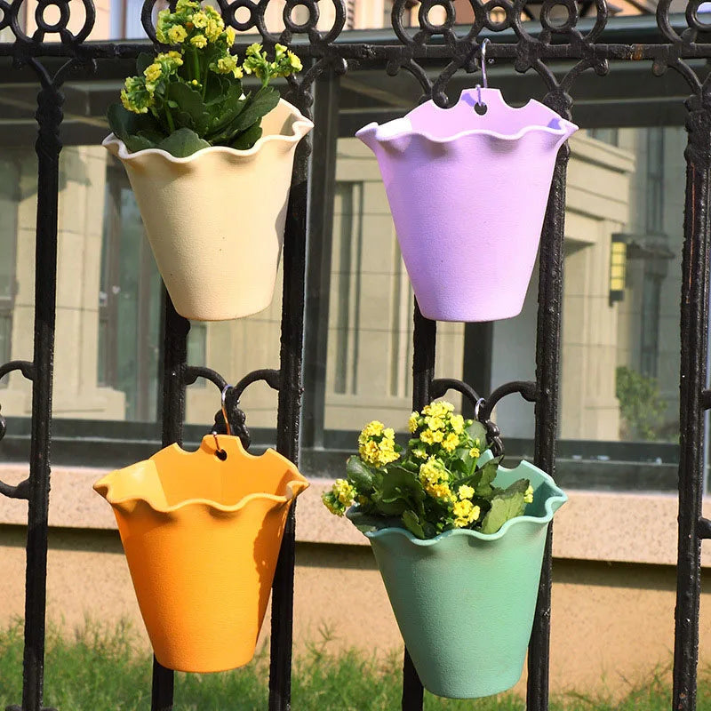 Four pastel-colored, ruffled plastic planters hang from a black metal fence, two containing small yellow flowers.