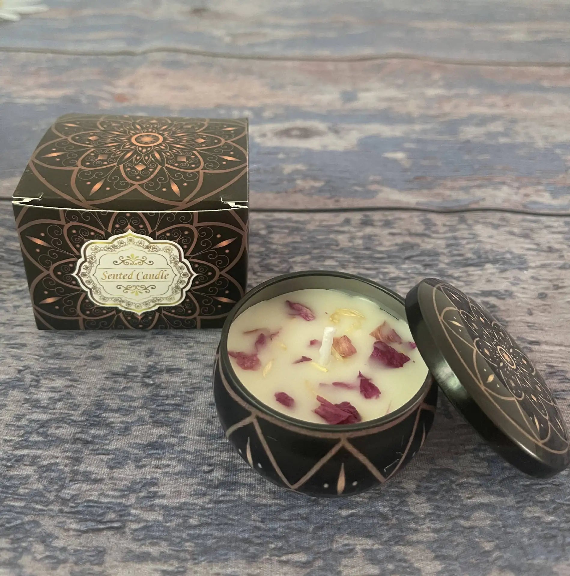Scented candle in a decorative black and bronze tin with matching box, topped with scattered dried floral petals.