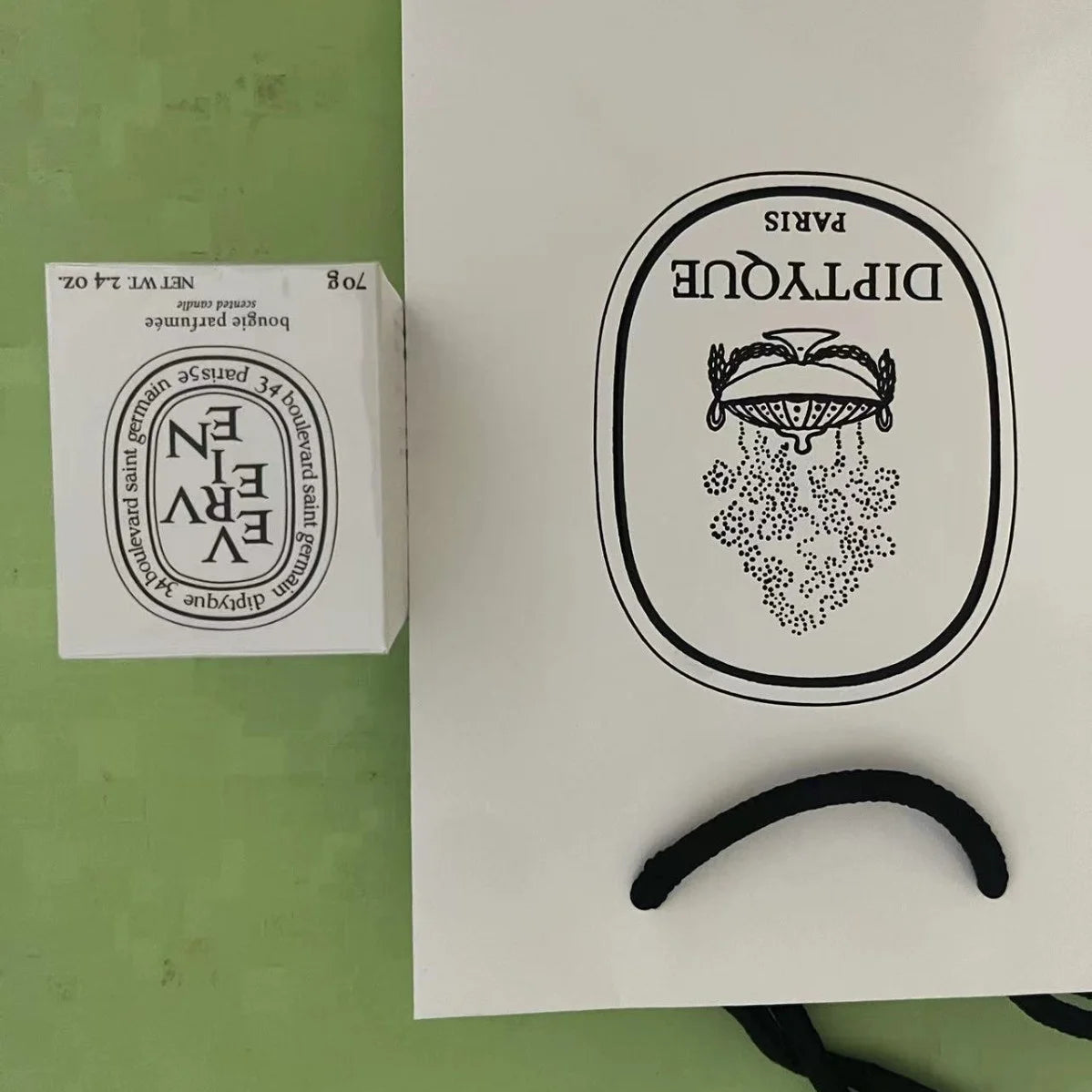 A luxury 70g scented candle in a white box next to a white paper gift bag with black branding and cord handles.
