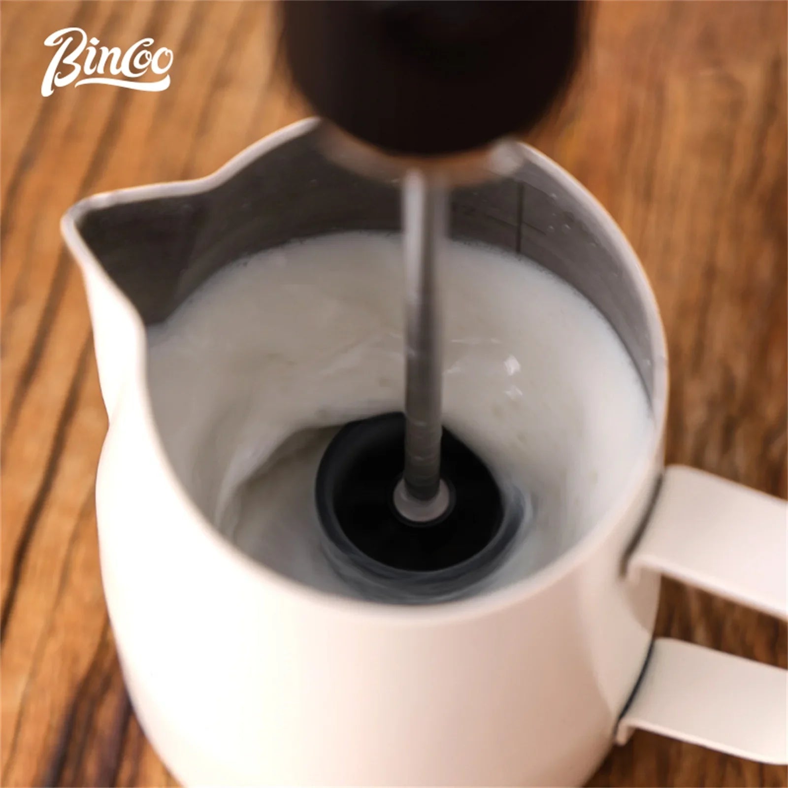 A handheld electric milk frother blends milk inside a matte white stainless steel pitcher on a wooden surface.