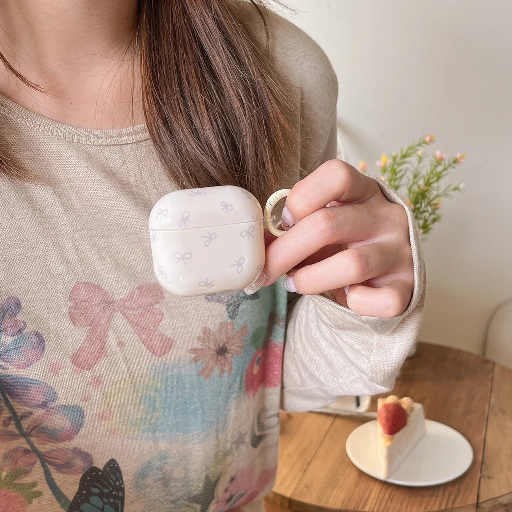 Cream AirPods case with grey bow print, smooth finish, featuring a side ring loop. Compatible with AirPods.