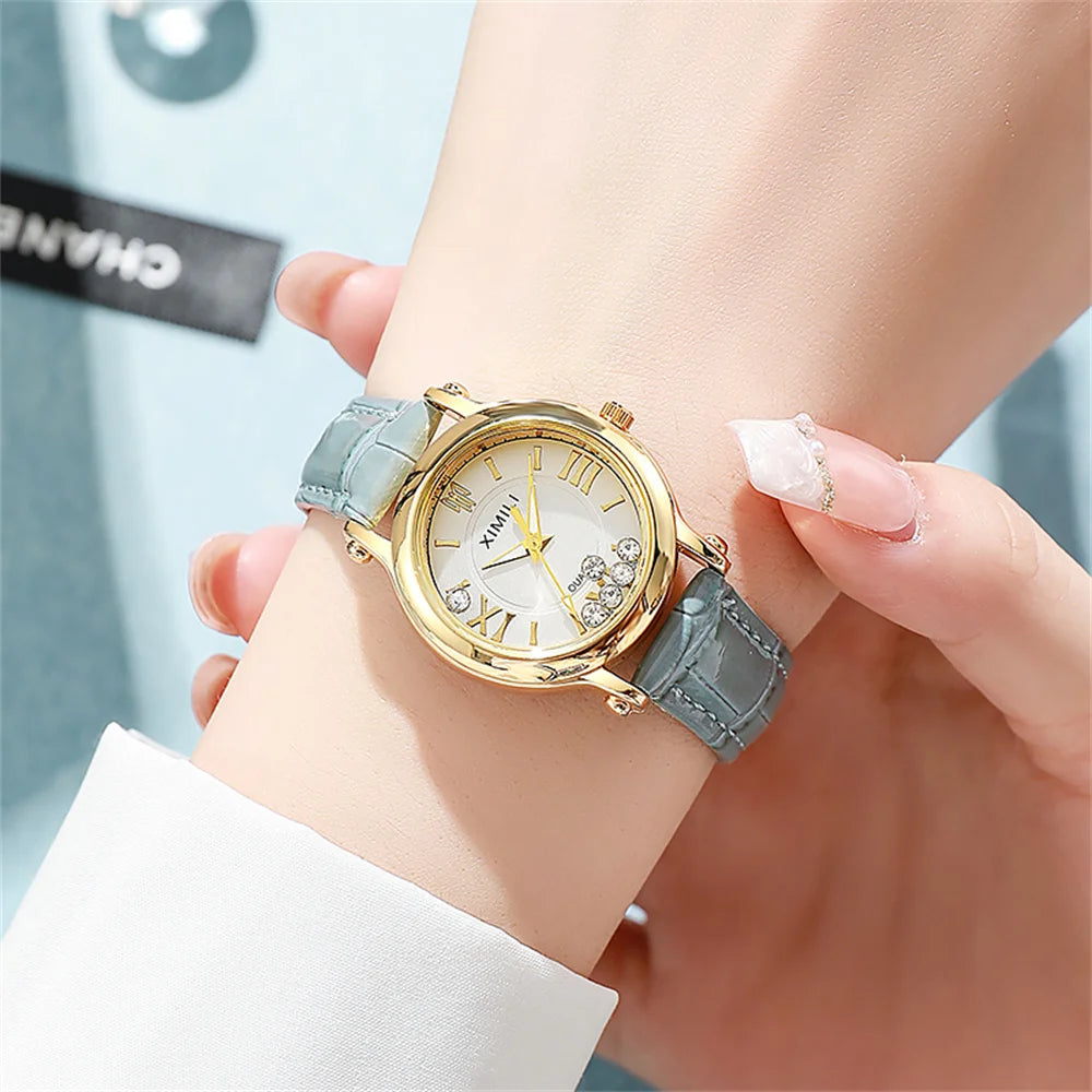 Women’s gold-tone quartz watch with a grey faux leather strap, white face, Roman numerals and decorative crystals.