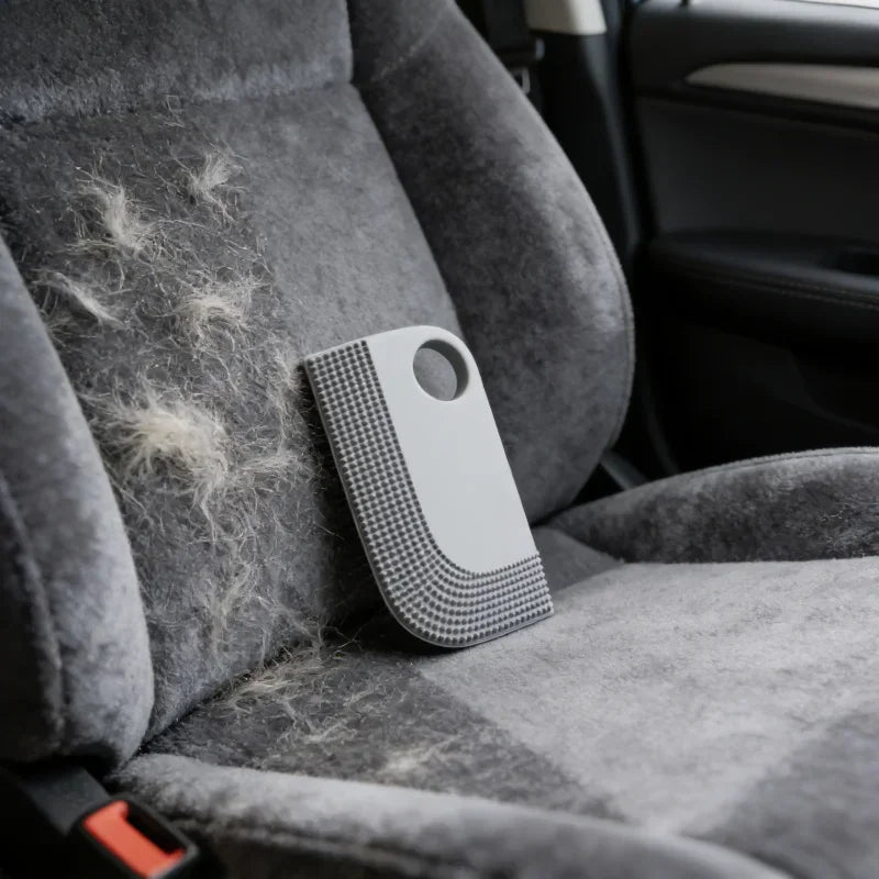 Grey rubber pet hair remover tool with textured edges resting on a car seat covered in pet fur.