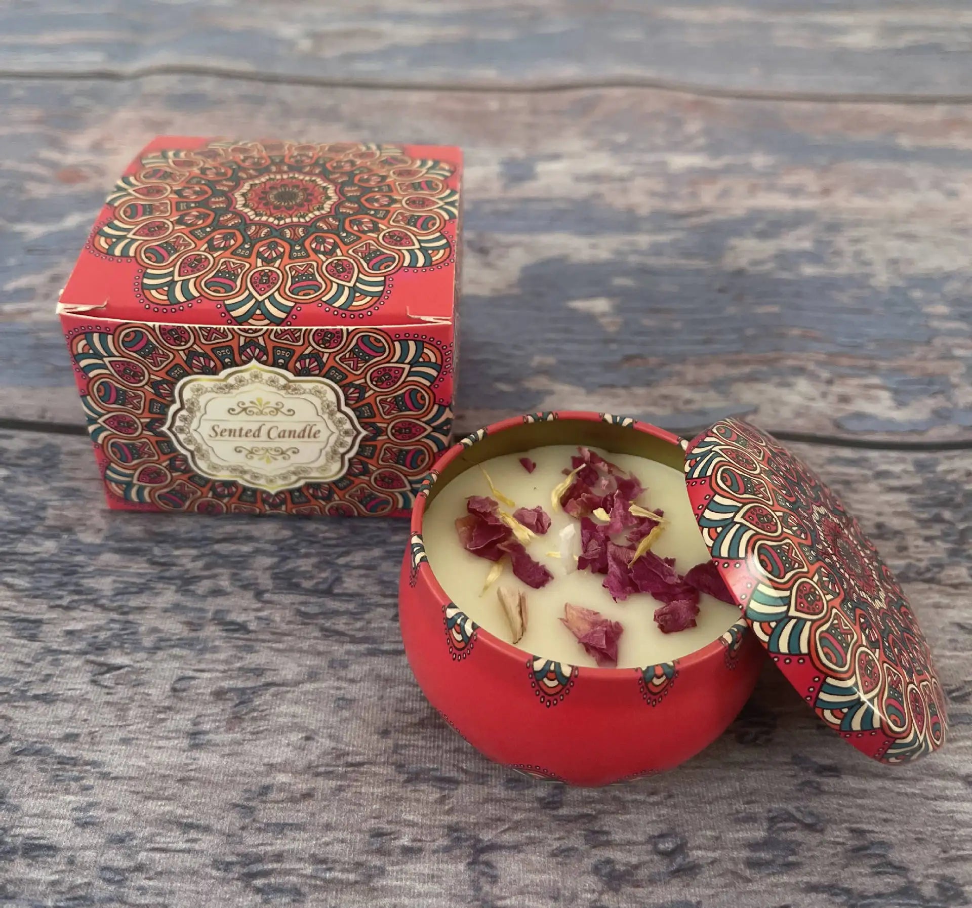 Red scented candle in a decorative mandala-print tin, topped with dried flower petals, next to its matching gift box.