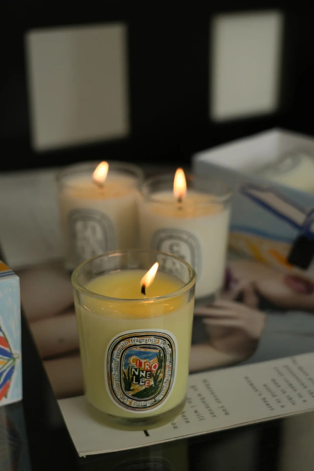 Three burning candles in clear glass jars with colourful oval labels on a reflective surface.