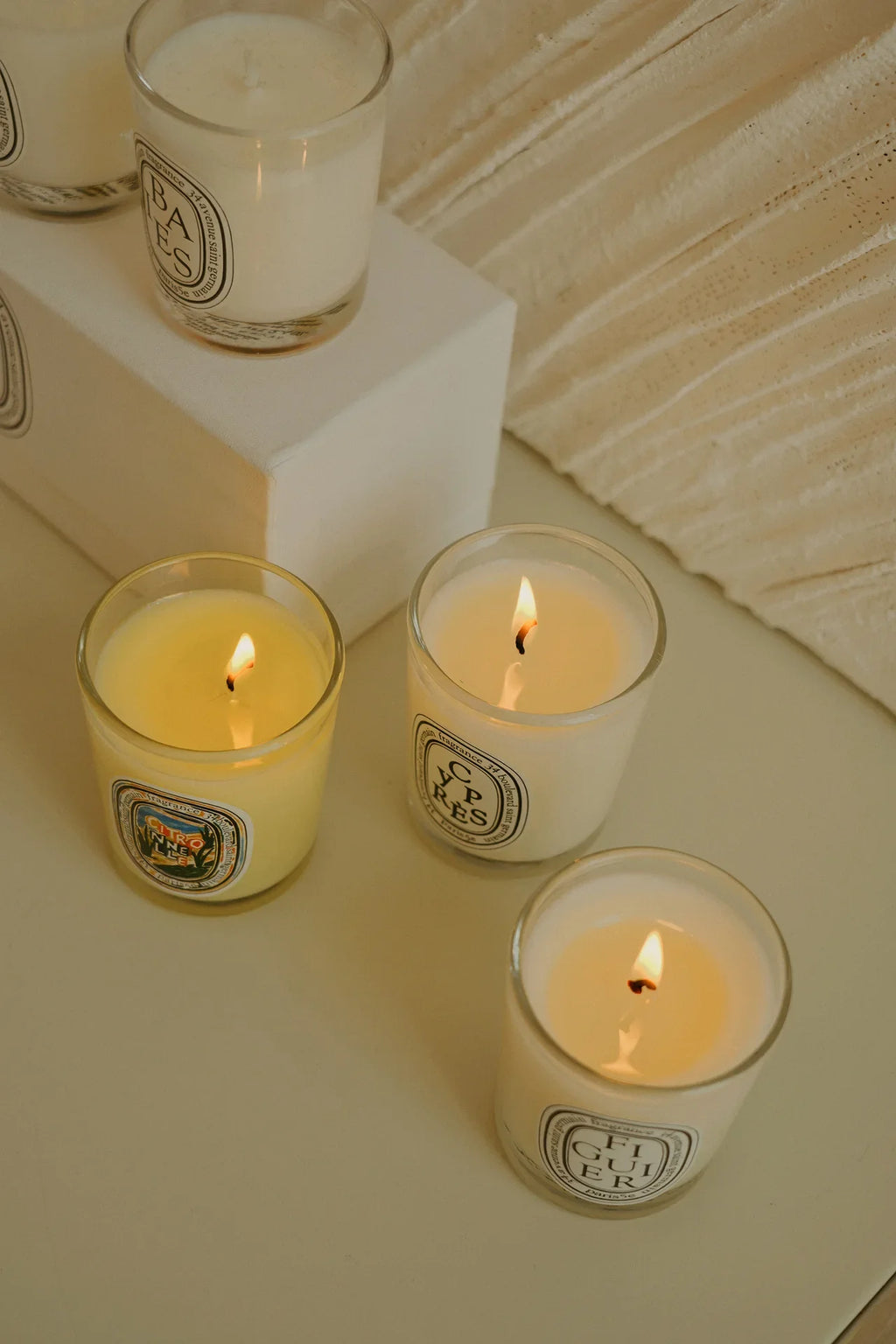 Four scented candles in glass jars, including three lit, arranged on a white surface with minimalist branding labels.
