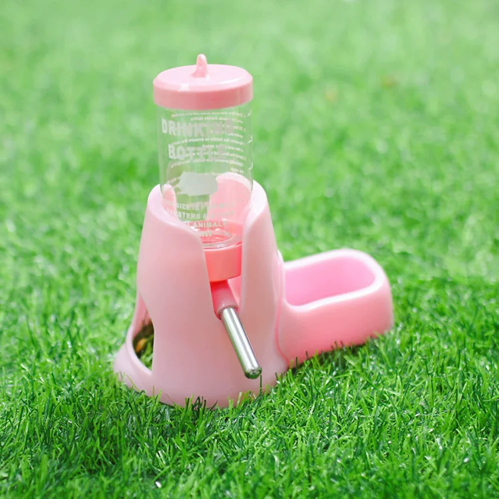 Pink plastic small animal water dispenser with a refillable bottle and integrated feeding bowl on a green lawn.